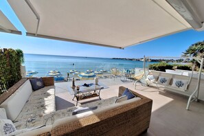 A sunlit outdoor seating area with wicker furniture, a glass table, and blue and white umbrellas. - Pearl of the Gulf Ortigia Holidays (Fontane Bianche)