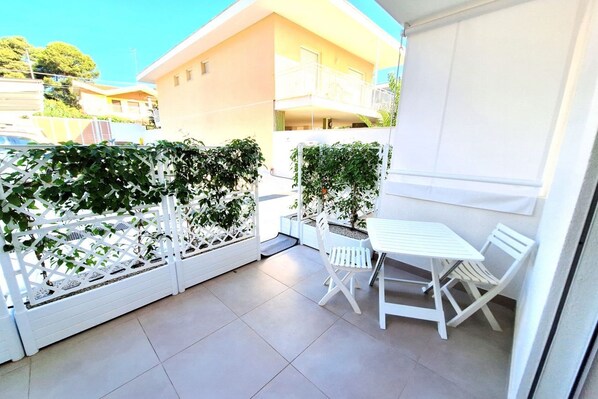 A balcony with a white table and chairs, enclosed by a white lattice fence, and a view of a building in the background. - Pearl of the Gulf Ortigia Holidays (Fontane Bianche)