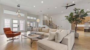 A spacious living room with a large beige sofa, a modern orange armchair, and a wooden coffee table. A large tree and a ceiling fan are visible in the room.
