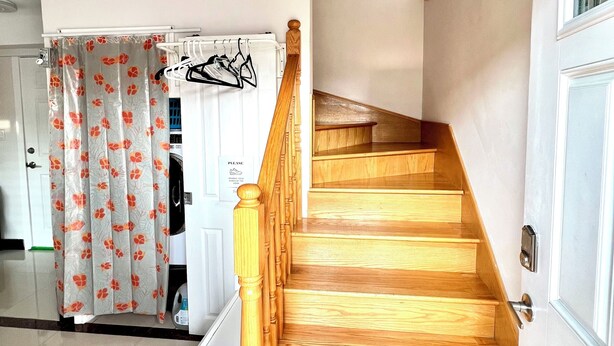 Un escalier en bois, doté d’une rampe, mène à un palier situé au deuxième étage. Une porte blanche se trouve sur la droite, ainsi qu’un mur blanc sur lequel est apposée une enseigne.