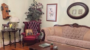 A traditional living room with a red armchair, a beige sofa, a wooden coffee table, a patterned rug, a small potted plant, a framed picture, and a wall-mounted mirror. - Charming 1-bedroom house in Historic Mineola, short walk to town (Mineola)