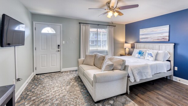 A bedroom with a bed, a sofa, a ceiling fan, a TV, and a window with curtains. - Walk to the Beach | 2 Pools | Military Welcome! (Biloxi)