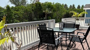 A wooden deck with a table and chairs, surrounded by a white railing and greenery.