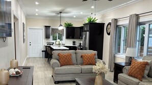 A spacious living room with a dark wooden coffee table, grey sofas with orange cushions, a ceiling fan, and a kitchen in the background.