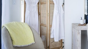 Um quarto com uma poltrona bege, uma pequena mesa redonda, um roupão branco em uma divisória de madeira e um espelho na parede.
