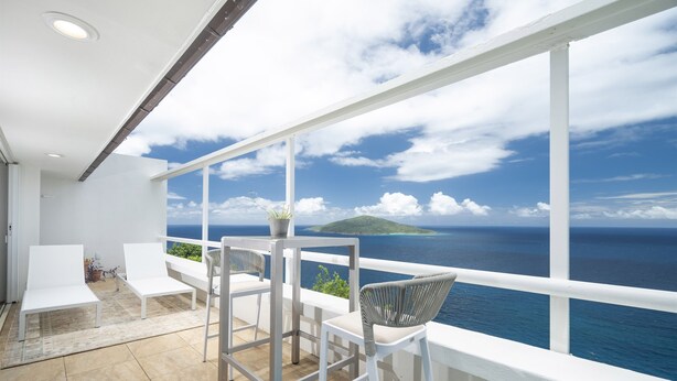 Varanda com cadeiras e mesa brancas, com vista para o mar e uma ilha distante.