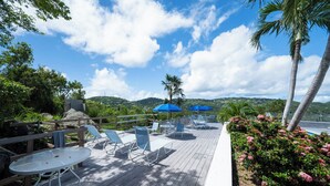 A wooden deck with lounge chairs and umbrellas, surrounded by lush greenery and a view of hills.