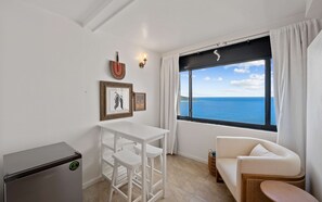 A compact room with a white table and chairs, a sofa, a small cabinet, and a window with a view of the sea.