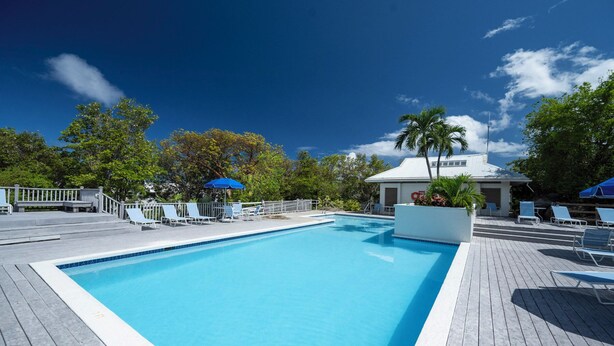 A rectangular swimming pool with a wooden deck surrounding it, lounge chairs, and a small building with a white roof.