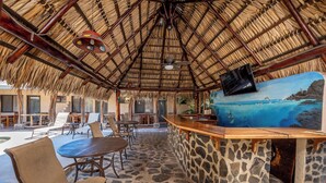 Un espace extérieur aménagé avec un toit de chaume, un sol en pierre, un bar en bois doté d’un comptoir en pierre et une fresque représentant un paysage côtier.