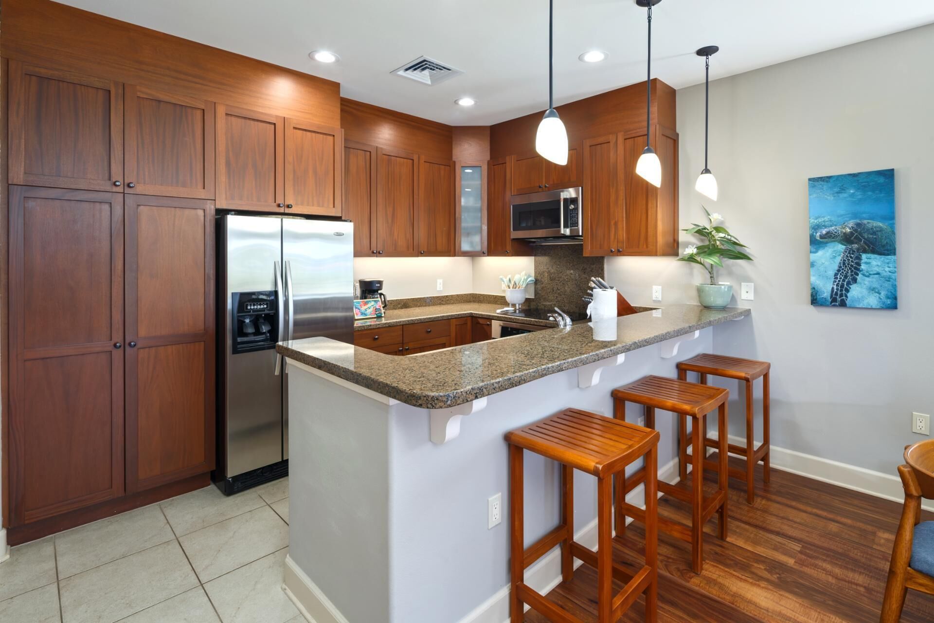 A modern kitchen with wooden cabinets, a stainless steel refrigerator, granite countertops, bar stools, and a painting of a sea turtle.