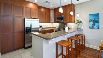 A modern kitchen with wooden cabinets, a stainless steel refrigerator, granite countertops, bar stools, and a painting of a sea turtle.