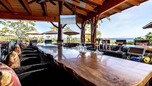 An outdoor bar area with wooden beams, a ceiling fan, and a view of the ocean. - Halii Kai Resort by KBM (Waikoloa)