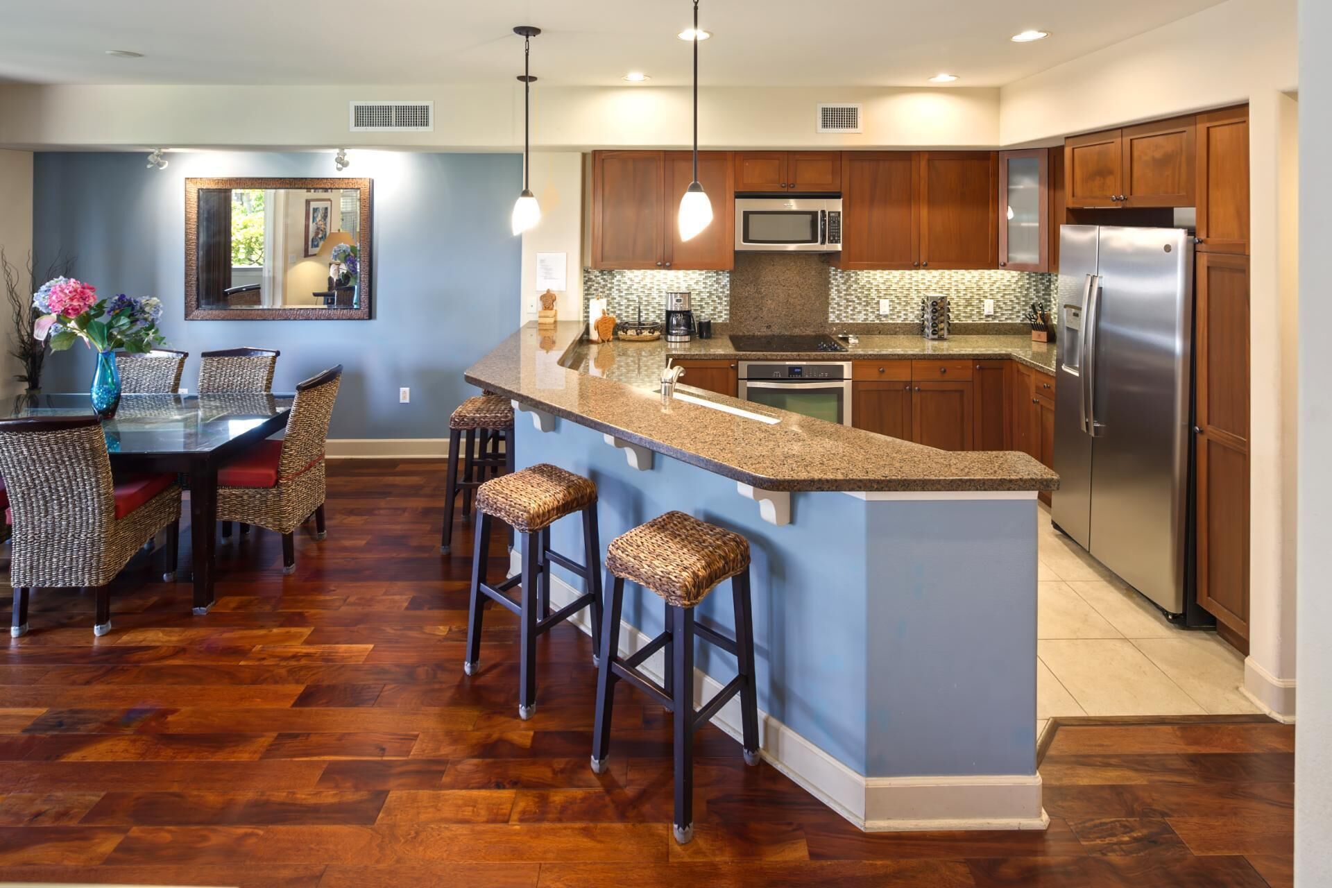 A modern kitchen with a central island, wooden cabinets, stainless steel appliances, and a dining area with wicker chairs and a glass table.