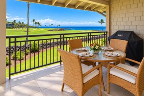 A covered outdoor dining area with wicker chairs and a table set for two, overlooking a golf course and the ocean. - Halii Kai Resort by KBM (Waikoloa)