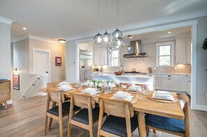 A modern kitchen with white cabinetry, a wooden dining table set for six, and pendant lighting. - Ocean-View Home- Chefs Kitchen,  Private Hot Tub, Dog Friendly, Double Patio (Pacific Beach)