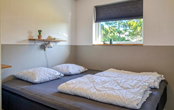 Una cama individual con ropa de cama blanca, una lámpara de noche, una ventana con vistas a árboles y una estantería con una planta y un libro.