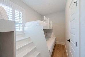 A compact room with a bunk bed, a window with blinds, a door, and a staircase. - Ocean Sunrise – Bayfront Luxury with Pool, Dock & Game Room. Steps to Gulf. Perfect for Families. (Bradenton Beach)