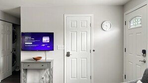 A modern living room with a flat-screen TV on a stand, a door, a clock, and a gray sofa. - Charming 2-bedroom at Issaquah Front St (Issaquah)