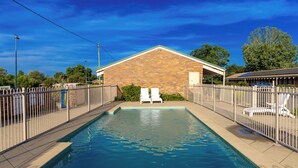 Une piscine rectangulaire, avec deux chaises longues placées en face, entourée d’une clôture, et un bâtiment avec un toit en pente en arrière-plan.