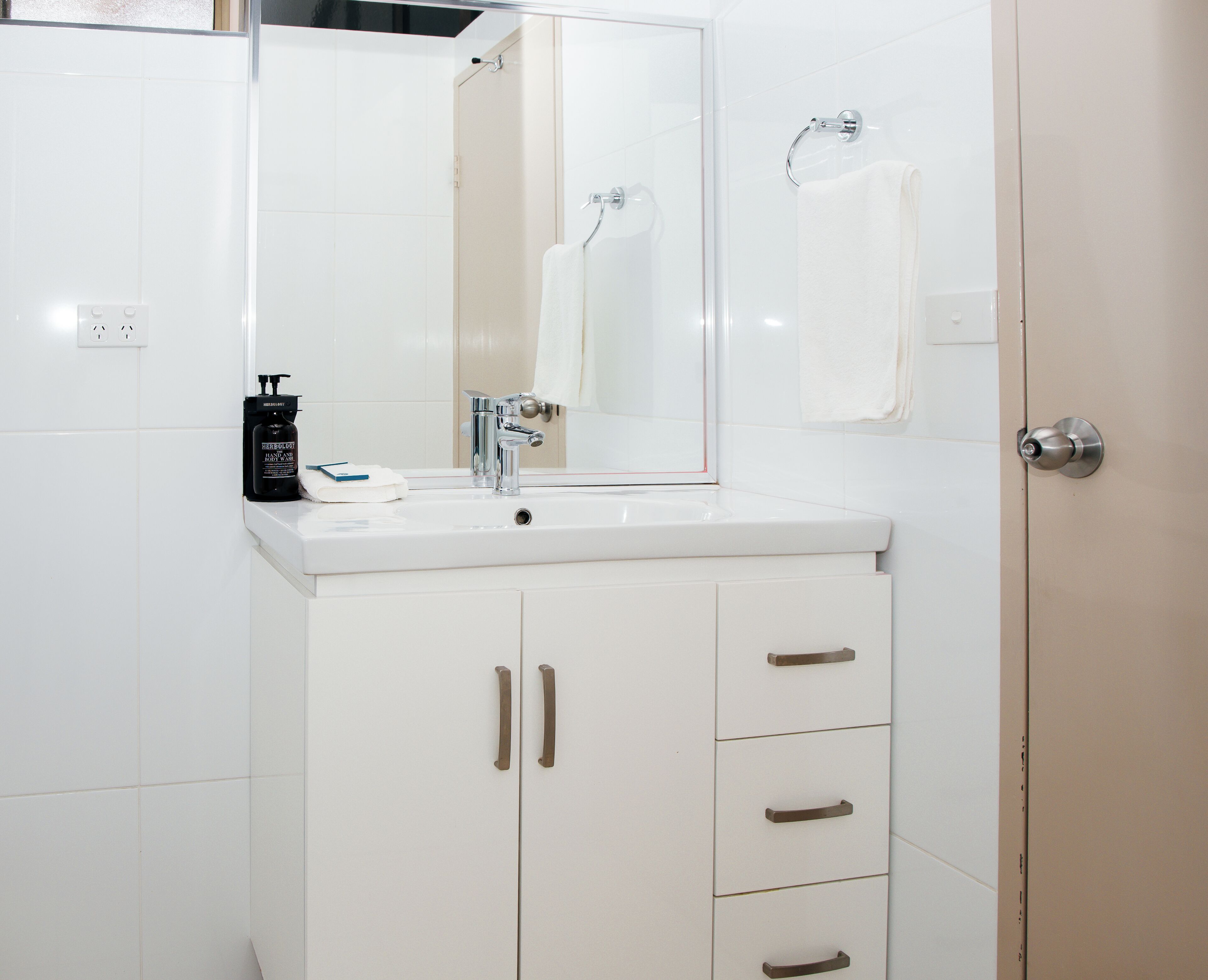 Un baño con lavamanos blanco, espejo, toallero y mueble con cajones.