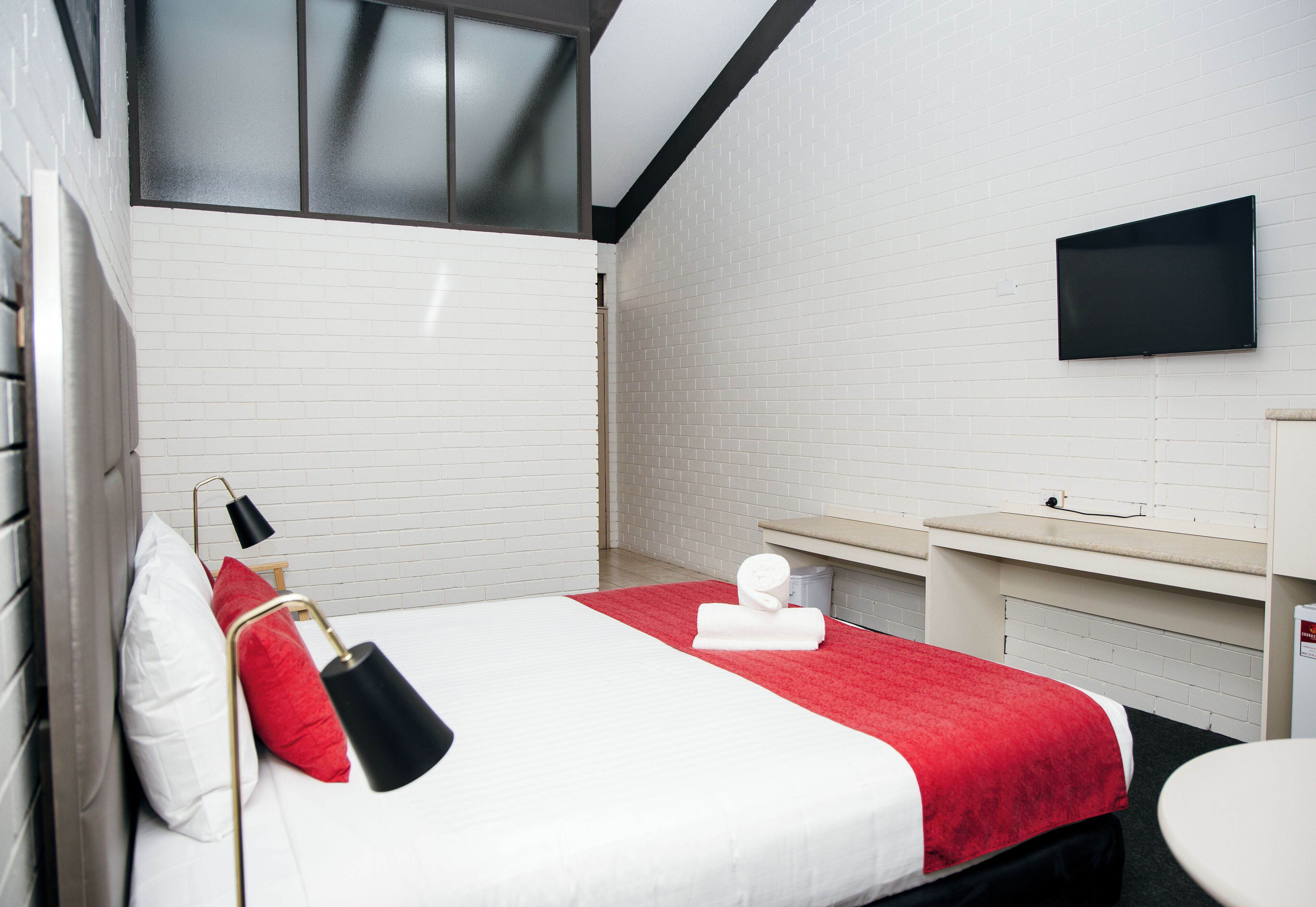 Una habitación de hotel con cama, televisor montado en la pared, una mesita y una ventana.