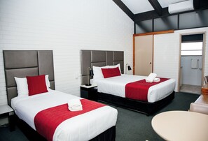 A hotel room with two beds, each with a red and white bedspread, a bedside table with a lamp, and a small round table.
