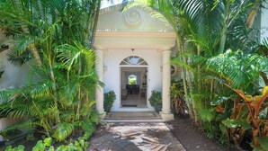 Jardí tropical que condueix a un edifici blanc amb un vestíbul cobert i un emblema circular a la part superior.
