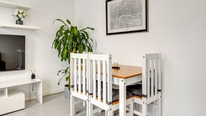 Un coin repas avec une table en bois et des chaises blanches, une plante en pot, une carte encadrée accrochée au mur et un meuble de rangement blanc avec un téléviseur.