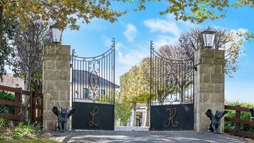 Stately entrance with ornate gates bearing the letter 'L', flanked by a tree and a wooden fence.