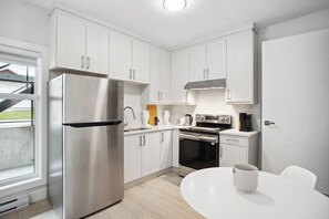 A modern kitchen with white cabinets, stainless steel appliances, a built-in oven, and a dining area with a white table and a cup. - Private 1BR Suite, New Build, 2 Guests, No Smoking (North Vancouver)