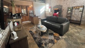 A spacious living area with a black leather sofa, a coffee table, a fireplace, and a kitchen in the background. - The Nest at Main Street Suites: Unique Apt in historic downtown Van Buren, AR (Van Buren)