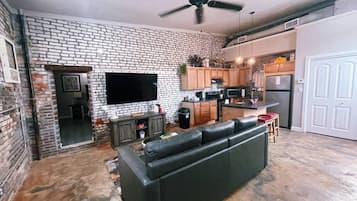 A living room with a black leather sofa, a flat-screen TV mounted on a brick wall, and a ceiling fan.