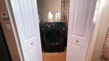 Un cuarto de lavandería con lavadora y secadora de carga frontal, dos botellas encima de la secadora y un termostato en la pared.