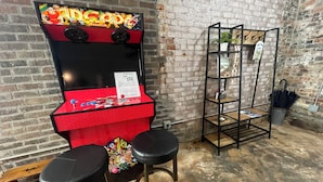 A retro arcade machine with two stools in front, a wooden shelf with plants, and a brick wall background. - The Nest at Main Street Suites: Unique Apt in historic downtown Van Buren, AR (Van Buren)