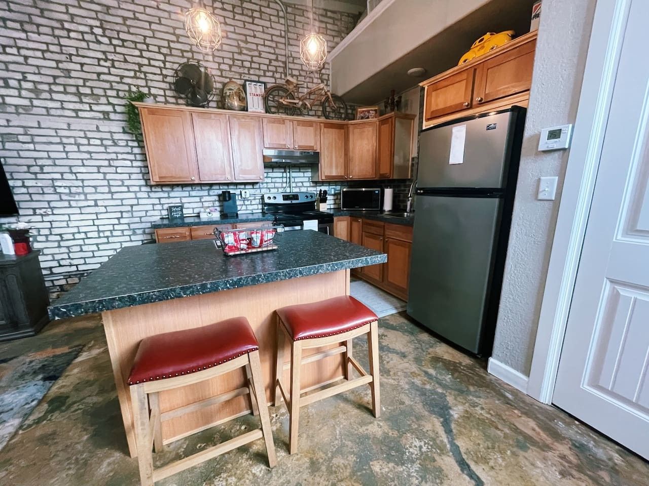 A kitchen with a brick wall, wooden cabinets, a granite countertop, a stainless steel refrigerator, and two red bar stools.
