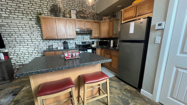 A kitchen with wooden cabinets, a granite countertop, a stainless steel refrigerator, and two red stools. - The Nest at Main Street Suites: Unique Apt in historic downtown Van Buren, AR (Van Buren)