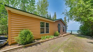 A wooden cabin with a window and a door, surrounded by greenery and a gravel path.