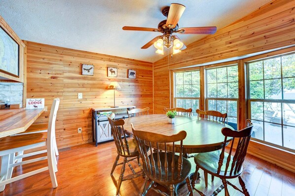 Un interior de madera con una mesa de comedor, sillas, un ventilador de techo y ventanas grandes.