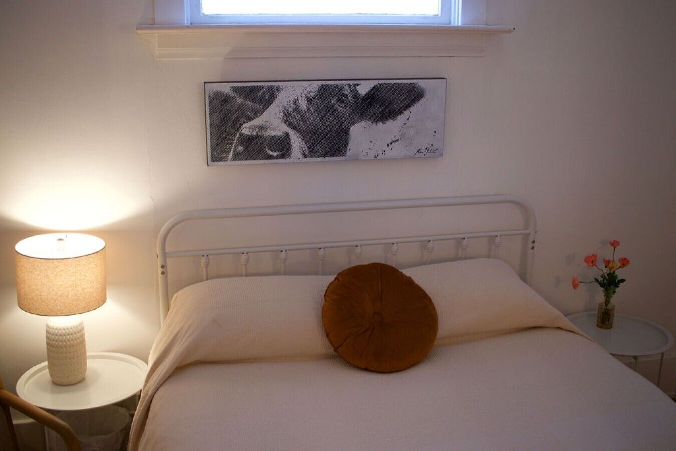 Een bed met een wit hoofdeinde, een rond kussen, een bedlampje, een klein tafeltje met een bloevas en een lijst aan de muur.