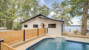 A modern house with a swimming pool, surrounded by trees and overlooking a body of water.