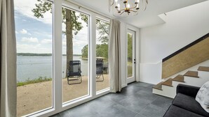 A spacious room with a large sliding glass door leading to a lakeside patio, a modern staircase, and a dark-colored sofa.