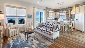 A spacious living room with a sofa, chairs, and a dining area. A kitchen with white cabinets and a view of the ocean is visible. - Oak Rest Barn, is a custom built gulf front home w/pool, pet friendly, $250 credit for beach gear! (Port Saint Joe)