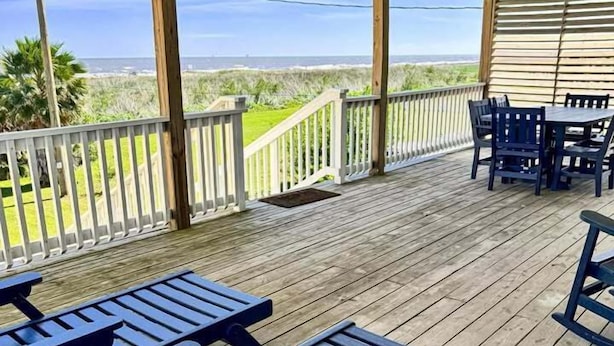 Une terrasse extérieure couverte, dotée d’un plancher en bois, de tables de pique-nique et de chaises, et offrant une vue sur une plage et l’océan.