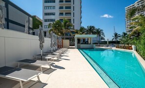 A pool area with lounge chairs and umbrellas, a small pavilion, and a tall building in the background.