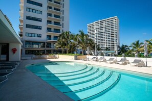 A swimming pool with lounge chairs and palm trees, surrounded by modern buildings.