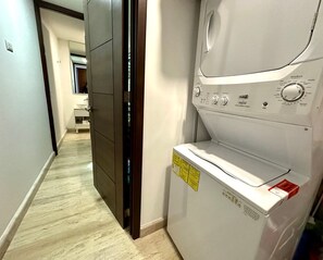 A laundry room with a washing machine and dryer, a door leading to another room, and a hallway with a light-colored floor.