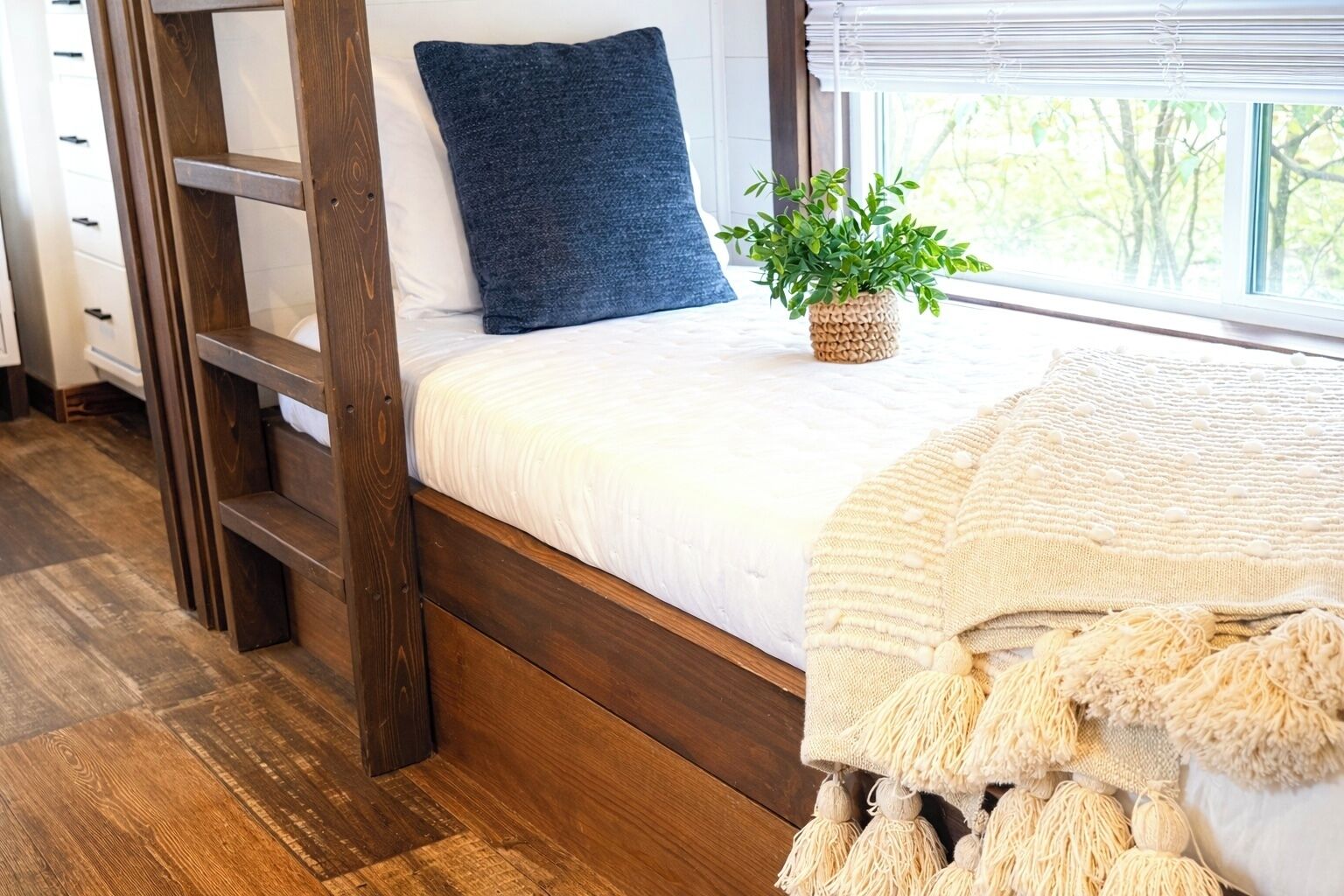 A bunk bed with a blue pillow, a plant on the bed, and a window with a view of trees.
