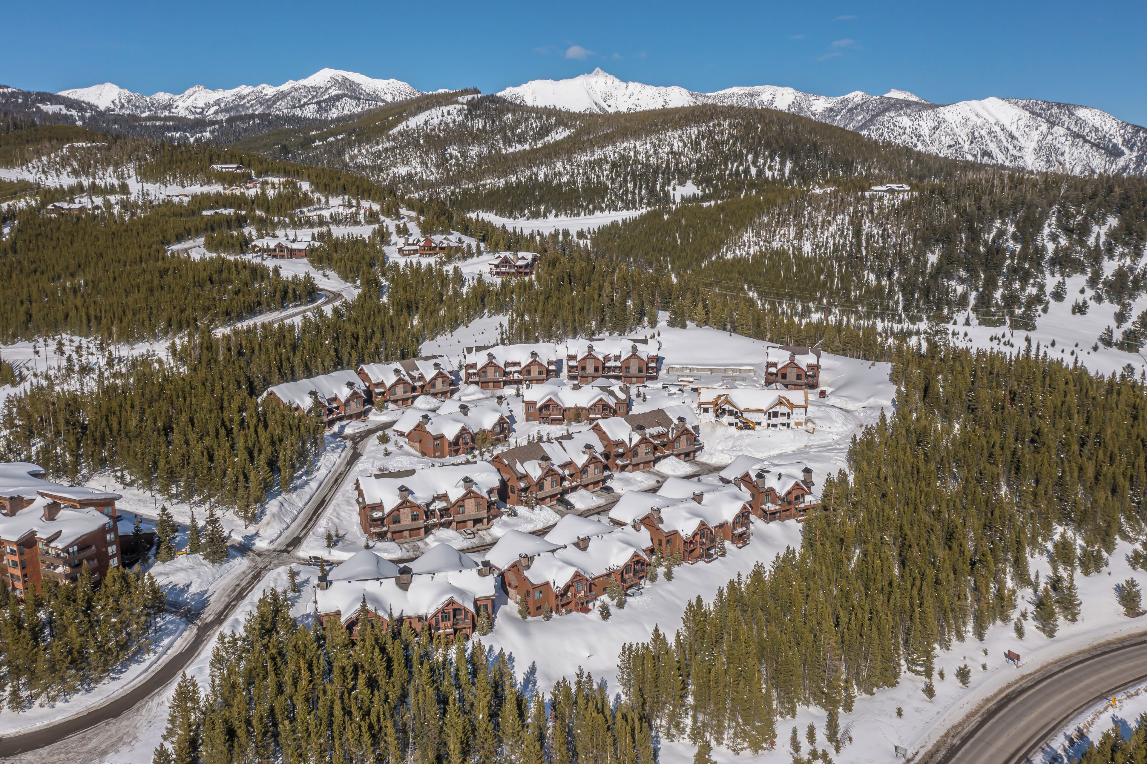 Un chalet enneigé composé de plusieurs bâtiments, entouré d’une forêt dense et de montagnes.