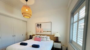 Une chambre à coucher comprenant un lit, deux tables de chevet, un luminaire suspendu, une fenêtre avec des stores et un tableau accroché au mur.
